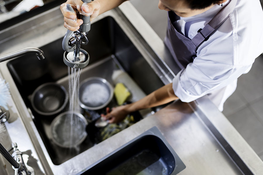 A restaurant employee using a faucet hose to spray food off of bowls in a stainless-steel commercial sink.